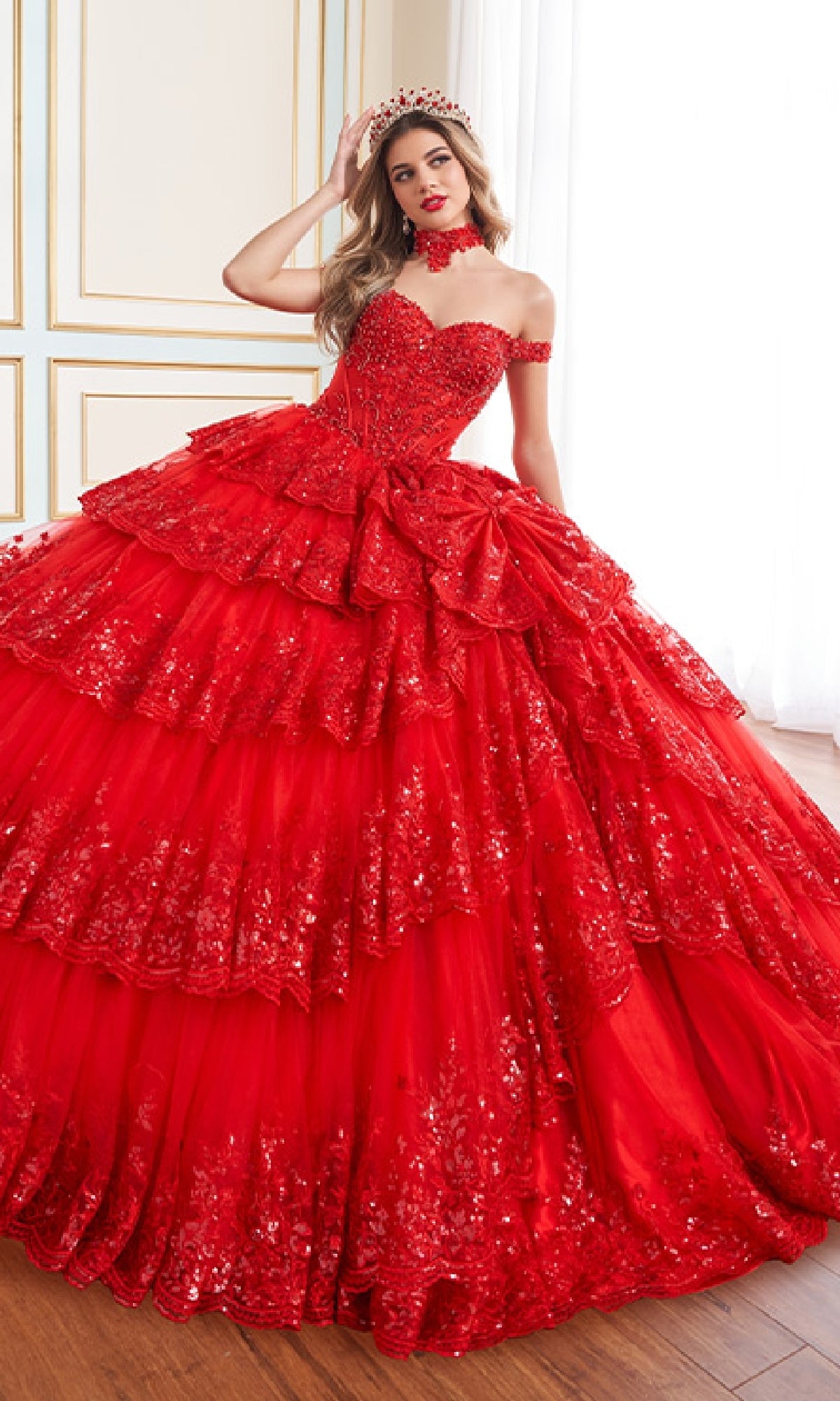 A woman stands indoors on a wooden floor wearing the Quinceanera Dress PR30181 by Princesa—a voluminous, off-the-shoulder red gown with layered ruffles and sparkling sequins.